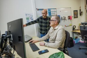 Collega achter computer en tweede persoon die iets aanwijst.